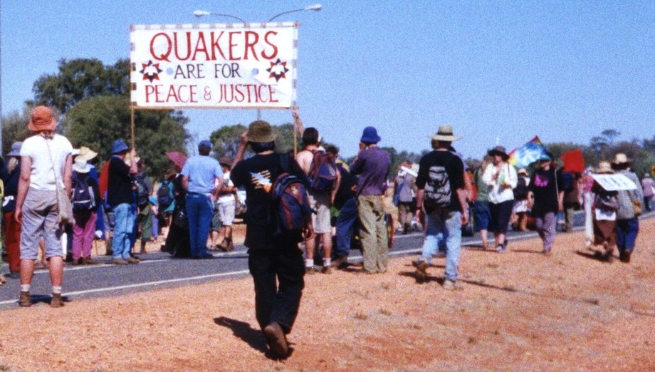 A group of people stand in and alongside the road leading to the U.S. military facility at Pine Gap, Australia. Some of them hold aloft a banner that says "Quakers Are For Peace & Justice."
