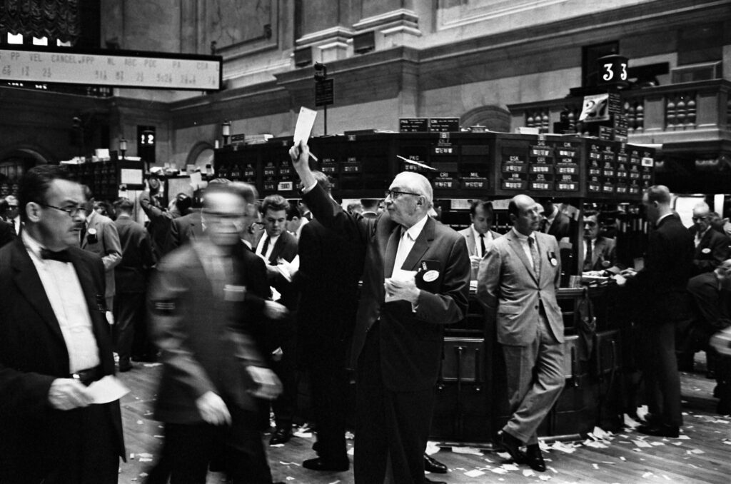 Corredores de bolsa ejerciendo su oficio en el suelo de la Bolsa de Nueva York a principios de la década de 1960. Son un grupo muy uniforme, de un tipo blanco, masculino y de mediana edad. 