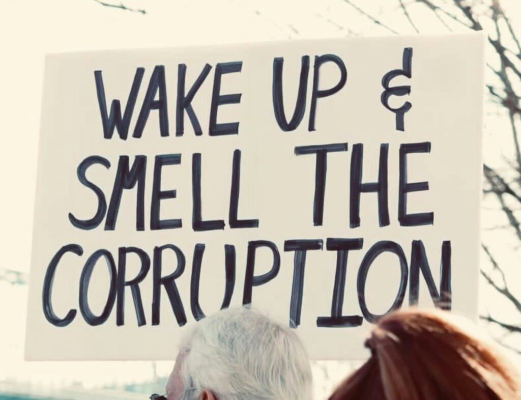 A closeup of a handmade sign that reads "WAKE UP & SMELL THE CORRUPTION." It is being held up in an outdoor setting, possibly by a white-haired person, the top of whose head is in the foreground.