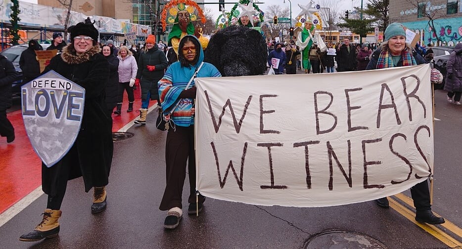 Two protestors marching down a Minneapolis street carry a white sheet upon which is written WE BEAR WITNESS. Next to them, another protestor carries a homemade sign in the shape of a shield bearing the legend DEFEND LOVE.