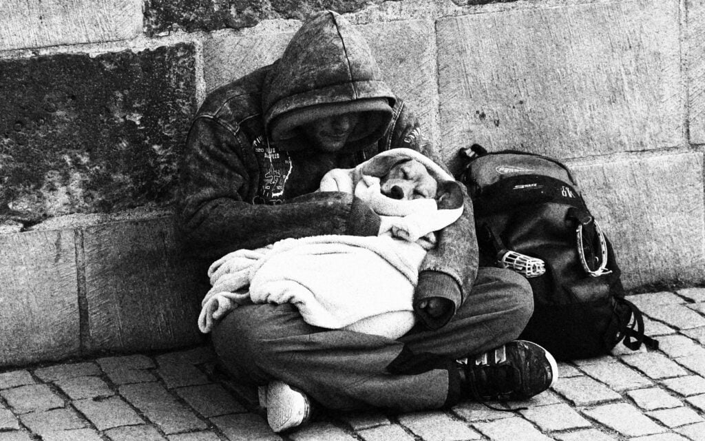 A man sits on a brick sidewalk, his back against the wall of a building. It's cold: He's wearing a hooded heavy jacket, his hands tucked inside his sleeves, and in his lap a dog sleeps, wrapped in a blanket.