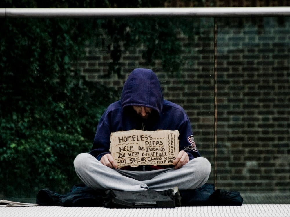 A man sits cross-legged on a sidewalk in front of a brick all, wearing gray sweatpants and a blue hooded sweatshirt. He holds a cardboard sign that says "HOMELESS... PLEAS HELP AS I WOULD BE VERY GREATFULL ANY SPEAR CHANG WOULD HELP ME, THANK YOU."