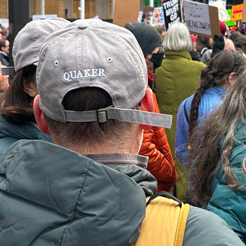 A photograph of the back of a man's head. He is wearing a gray baseball cap that says QUAKER in small white letters across the back. Around him we can see a crowd of protestors, some carrying signs criticizing the Trump administration and the invasion of Venezuela.
