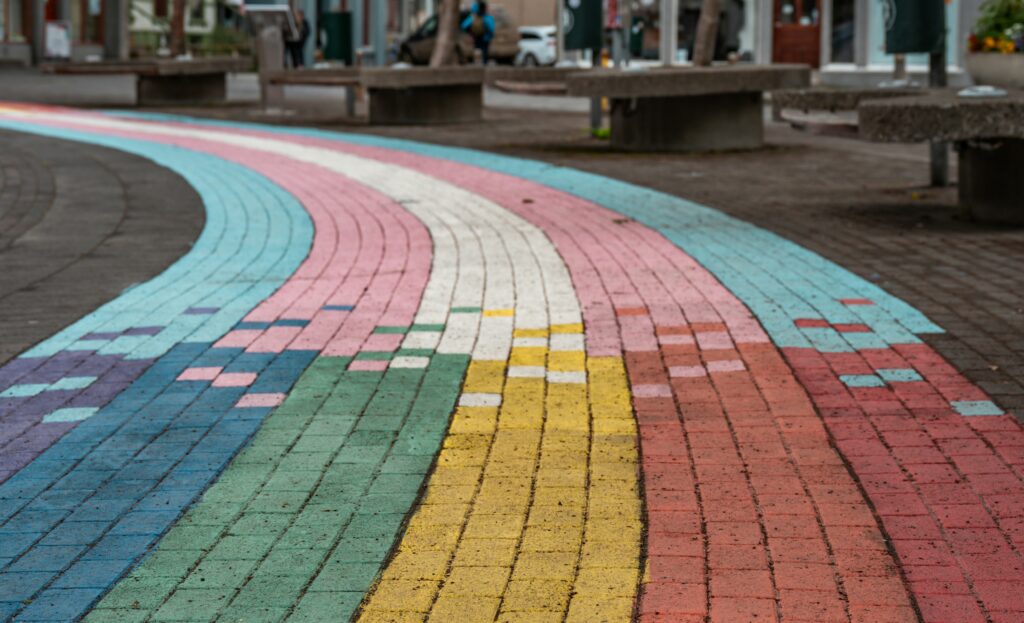A photograph of a city square, paved with bricks. Some of the bricks have been painted to form a rainbow path, but midway down the path, the colors change to those of the trans pride flag.