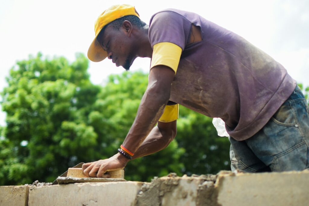 A Black man in jeans, a purple T-shirt over a yellow T-shirt, and a yellow baseball cap, uses a trowel to spread mortar across the top of a stone in a wall.