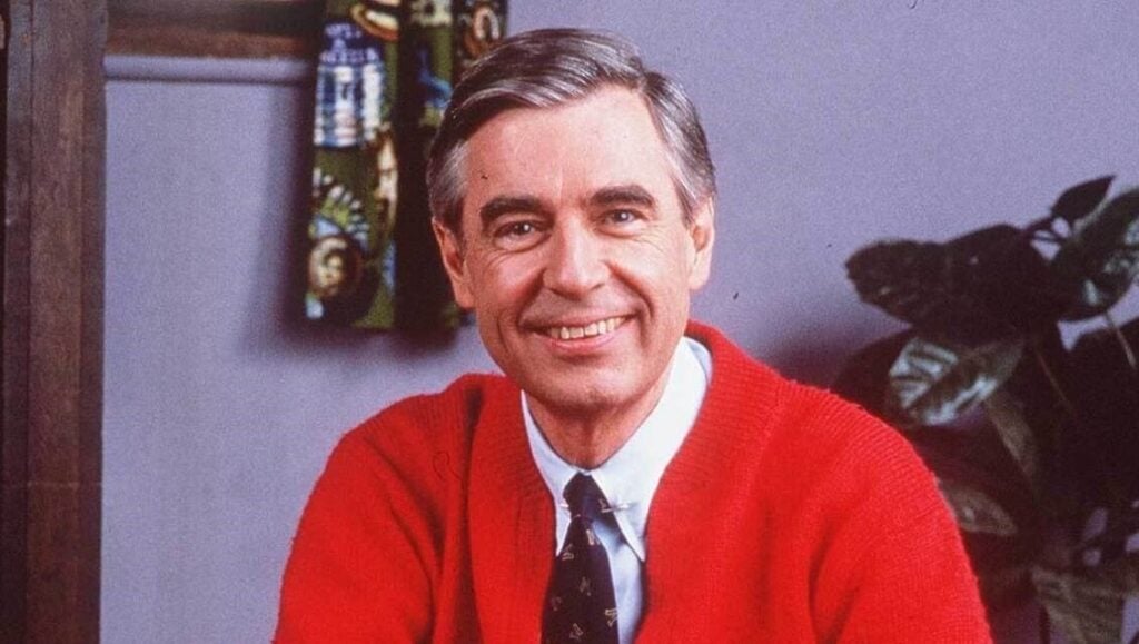 A color photograph of a smiling Fred Rogers on the set of Mister Rogers Neighborhood, sitting on the bench by the front door in a red cardigan sweater.