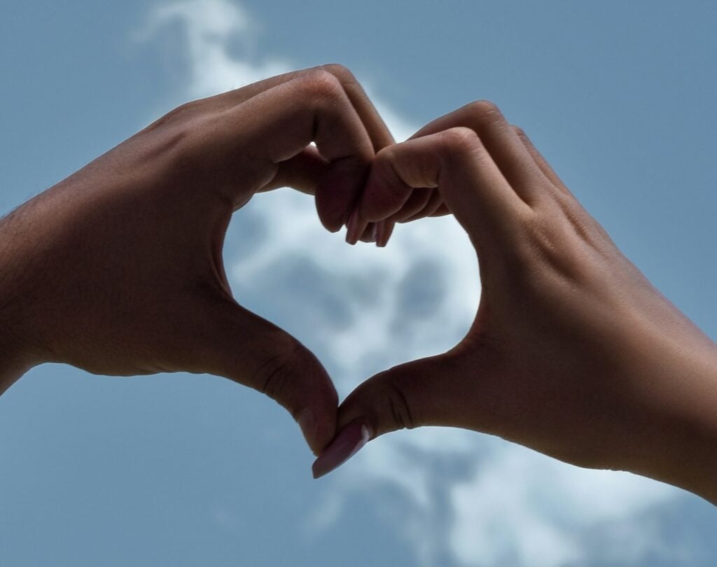 A pair of dark-skinned hands held up against a white cloud in a blue sky, forming the shape of a heart icon.