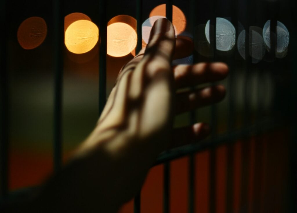A photograph of a forearm, stretched out in front of the bars of a jail cell, with blurry multicolored lights in the background.