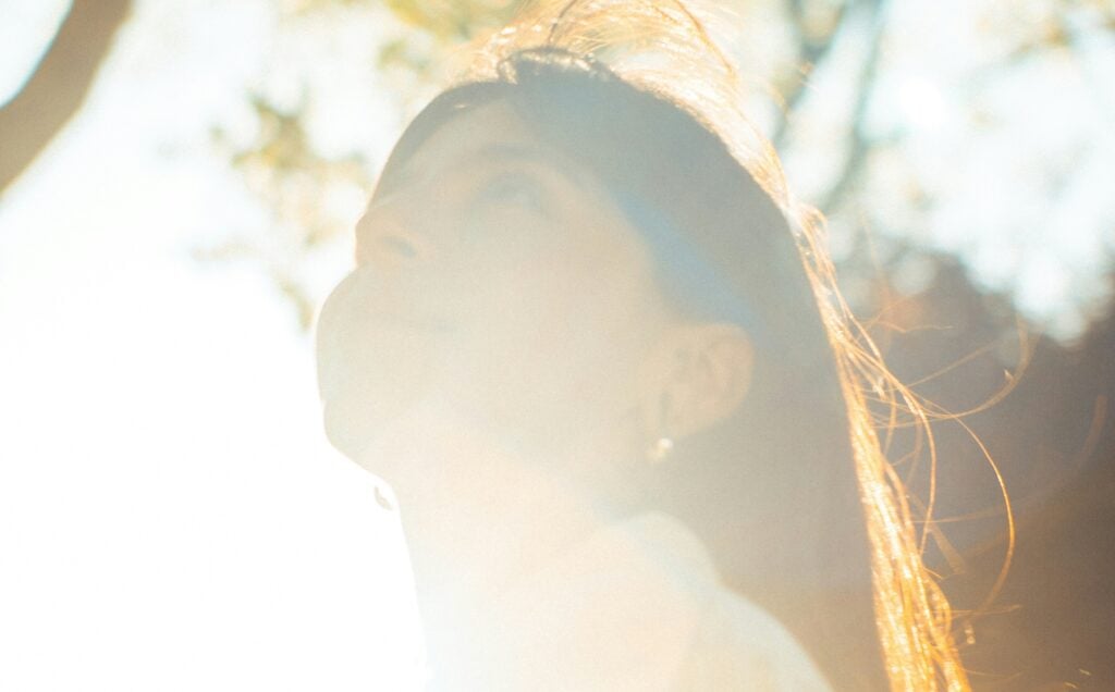 A photograph of a young woman with long, dark hair standing amid some trees. She looks up to the sky and the sun shines brightly upon her.
