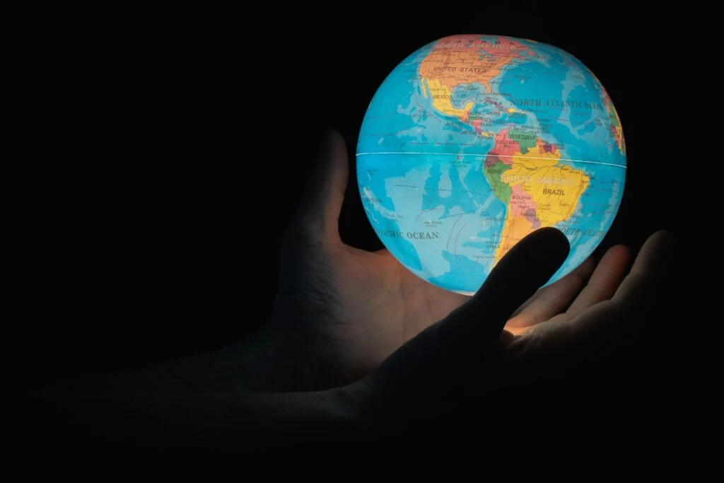 A photograph of a small globe of the planet Earth, illuminated from its interior, being cradled in a pair of human hands in the darkness.