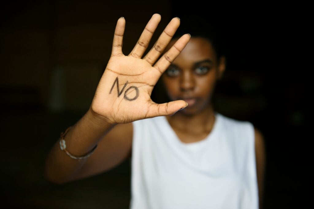 A photograph of a young Black person in a white sleeveless shirt, facing the camera. They have raised their right hand, and on the palm they have written the word NO in large black letters.