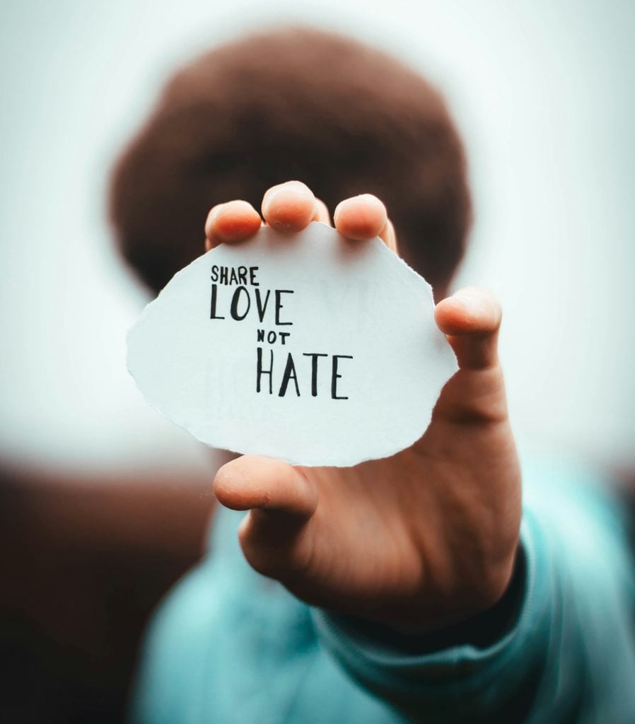 A photograph of a Black person holding their hand out so as to obscure their face. In their hand, they hold a piece of white cardboard on which the message SHARE LOVE NOT HATE has been printed.