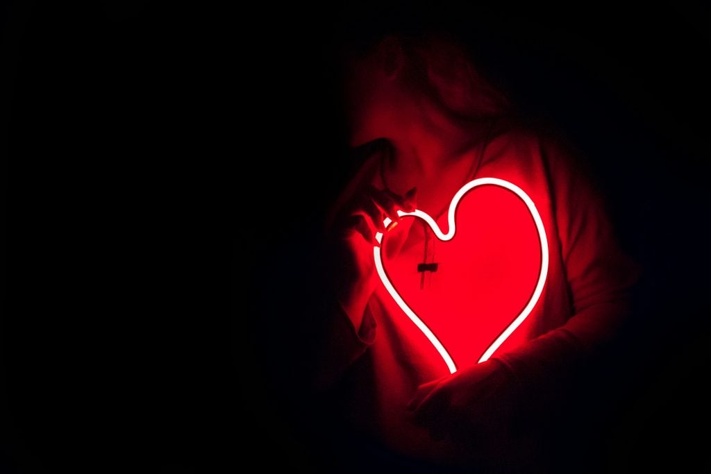 A photograph of a person standing in darkness, clutching to their chest a glowing neon sculpture of a cartoon heart.