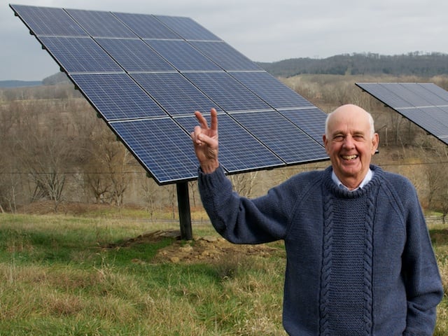 Wendell Berry, an older White Man balding on top with white hair at the temples, stands in front of a solar energy panel in a field, raising his right arm and making a peace sign. He wears a blue sweater.