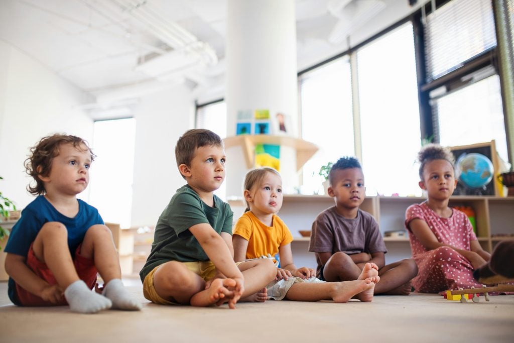 A photograph of five very young Black and White children sitting on the floor of a nursery or a preschool classroom.