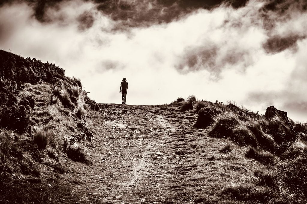 A lone person wearing a broad-brimmed hat walks up an inclined trail toward a cloud-filled sky.